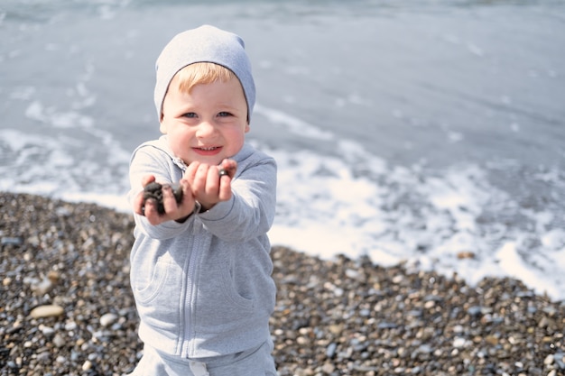 Kind blonder Junge spielt mit Felsen und Sand am Strand an einem sonnigen Tag im Frühjahr oder Herbst.