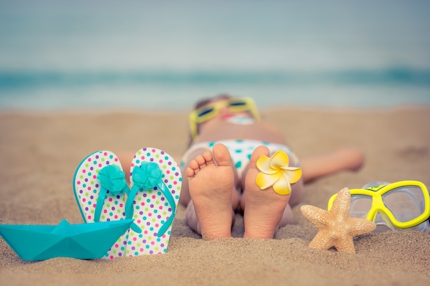 Kind, das am Strand mit einer Blume am Fuß und Flipflops und einem Seestern auf dem Sand liegt