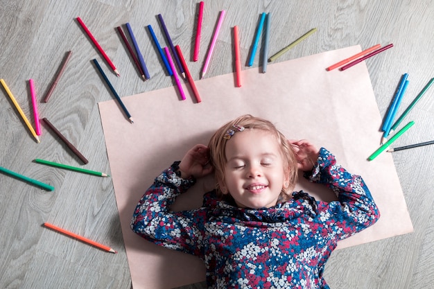 Kind, das auf dem Boden auf Papier mit geschlossenen Augen nahe Buntstiften liegt. Kleines Mädchen malt, zeichnet. Draufsicht. Kreativitätskonzept.
