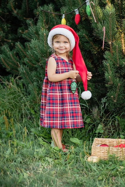 Kind, das auf ein Weihnachten im Holz in Juli wartet. Porträt von kleinen Kindern in der Nähe von Weihnachtsbaum. Mädchen schmücken Weihnachtsbaum mit Kugeln.