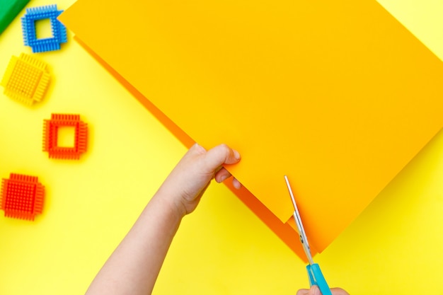 Kind, das farbiges orange Papier mit einer Schere auf einem Tisch für etwas Handwerk schneidet