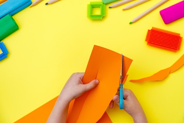 Kind, das farbiges orange Papier mit einer Schere auf einem Tisch für etwas Handwerk schneidet