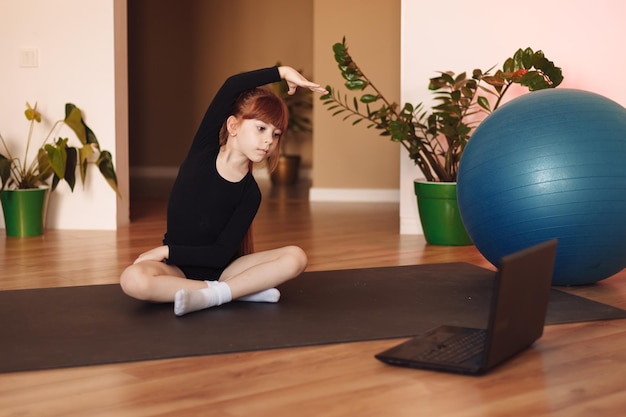 Kind das Mädchen sitzt vor dem Laptop auf der Sportmatte und wiederholt die Übungen online