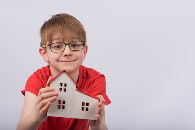 Kind, das Modell des Hauses auf weißem Raum hält. Netter Junge mit Brille, die Spielzeughaus hält. Homeschooling