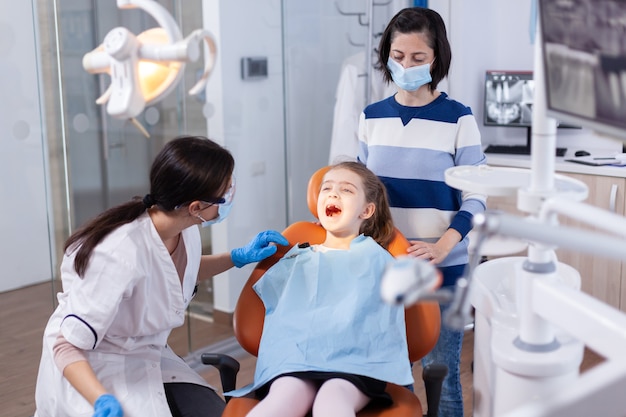 Kind, das während der mündlichen Prüfung den Mund offen hält, sitzt auf dem Zahnarztstuhl. Zahnmediziner während der Kinderhöhlenberatung in der Stomatologie-Büro mit moderner Technologie.