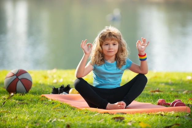 Kind, das Yoga-Asana auf Rollmatte im Park meditiert, scherzt Mädchen, das Yoga im Freien praktiziert, Kind, Junge