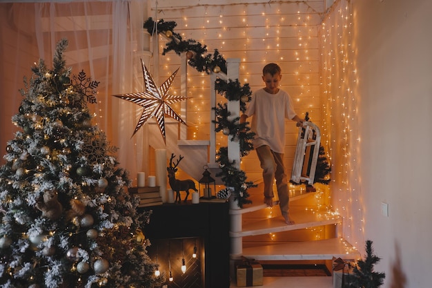 Kind, das zu Hause Weihnachtsbaum schmückt. Kleiner Junge in Strickpullover mit Weihnachtsschmuck. Familie mit Kindern feiert Winterferien. Kinder dekorieren Wohnzimmer und Kamin zu Weihnachten.