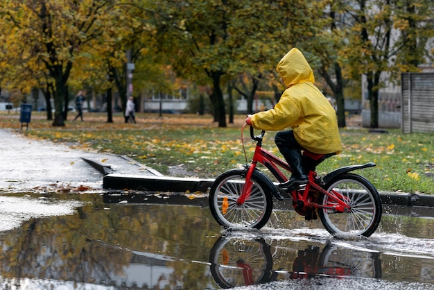 Kind fährt Fahrrad im Regen und Pfützen. Junge im gelben Regenmantel ist allein durch die Pfützen reiten.