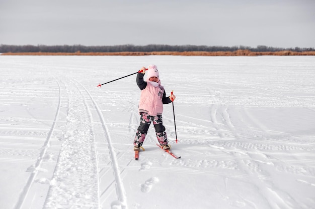 Kind fährt im Schnee Ski Fröhliches Mädchen im warmen Trainingsanzug lernt Skifahren am gefrorenen Flussufer und winkt mit der Hand Winterlandschaft schneebedeckter Hintergrund