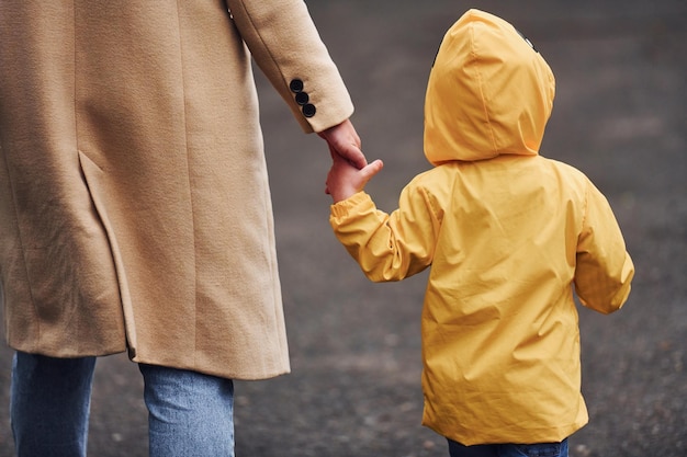 Kind im gelben wasserdichten Mantel, der die Hand der Mutter hält und im Freien spazieren geht