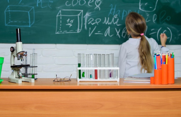 Kind in der Nähe der Schule Tafel Hintergrund defokussiert Schule Biologieunterricht Kind Studie Biologie mit Ausrüstung in der Schule Tag des Wissens Biologie-Konzept Mikroskop und Reagenzgläser auf dem Tisch im Klassenzimmer