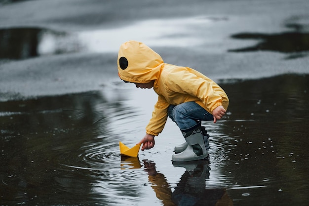 Kind in gelbem wasserdichtem Mantel und Stiefeln, das nach dem Regen mit handgefertigtem Bootsspielzeug aus Papier im Freien spielt