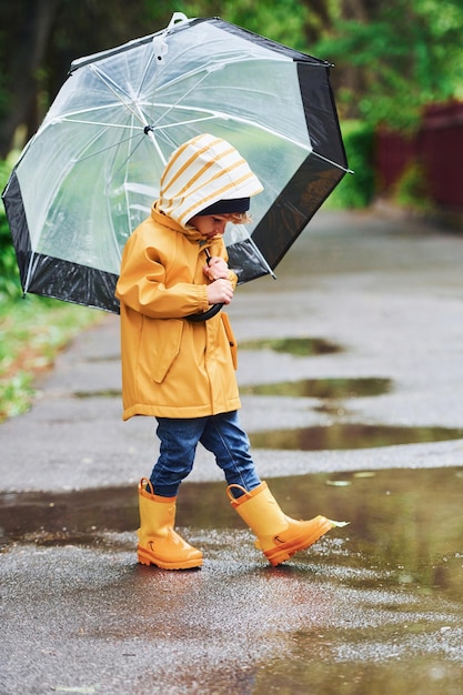 Kind in gelben, wasserdichten Umhangstiefeln und mit Regenschirm, der nach dem Regen im Freien spielt