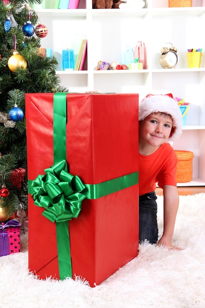 Kind in Weihnachtsmütze in der Nähe von Weihnachtsbaum mit großem Geschenk