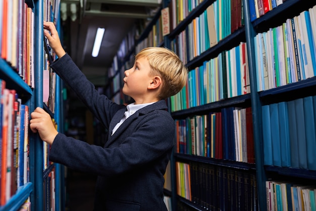 Kind Junge sucht und wählt Buch in Buchhandlung, steht in der Nähe von Regalen, wird lernen und lernen. Seitenansicht