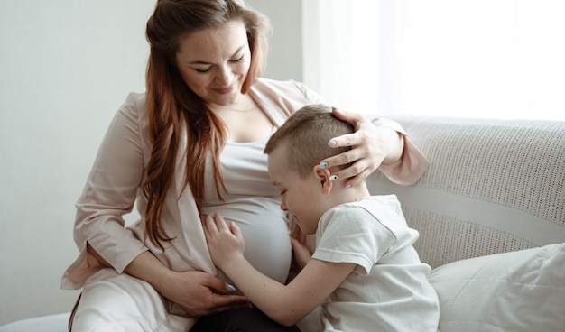 Kind Junge umarmt und küsst den Bauch der schwangeren Mutter auf der Couch zu Hause.