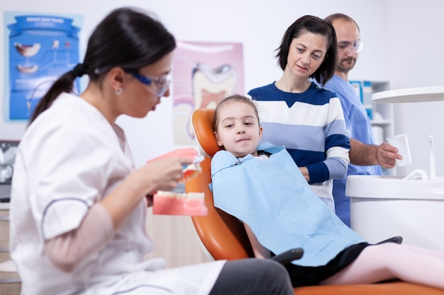 Kind lernt, wie man Zahnbürste von einem jungen Zahnarzt in Kleinem Mädchen und Mutter verwendet, die stomatolog über Zahnhygiene in der Zahnklinik mit Kiefermodell sprechen.