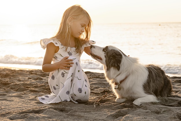 Kind Mädchen im Sommerkleid spielen mit Hund Haustier am Strand bei Sonnenaufgang am Morgen