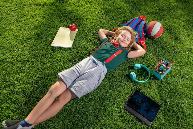 Kind mit Buch und Taplet und ruht sich nach der Schule auf dem Gras aus. Glücklicher Junge, der sich auf dem Gras entspannt