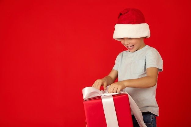Kind mit roter Weihnachtsmütze löst neugierig ein weißes Band an einer großen Geschenkbox