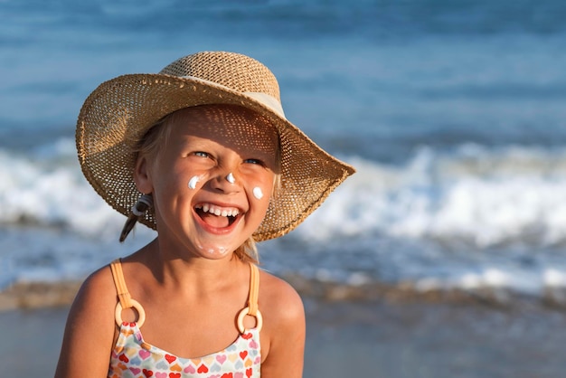 Kind mit Sonnencreme im Gesicht lächelt Kleines Mädchen in einem Hut auf dem Hintergrund des blauen Meeres