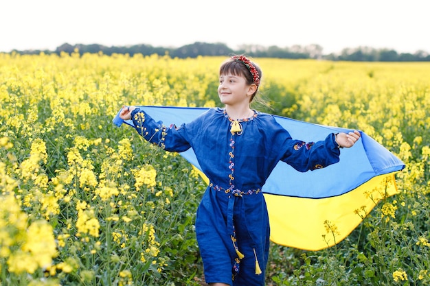 Kind mit ukrainischer Flagge im Rapsfeld Ein Mädchen in einem bestickten Hemd läuft mit der ukrainischen Flagge in den Händen über das Feld