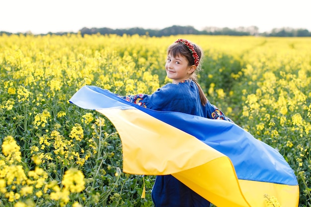 Kind mit ukrainischer Flagge im Rapsfeld Ein Mädchen in einem bestickten Hemd läuft mit der ukrainischen Flagge in den Händen über das Feld