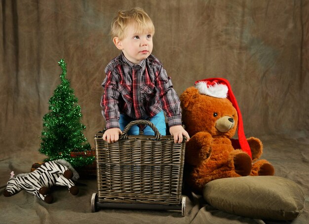 Kind sitzt am Korb in der Nähe von Weihnachtsbaum und riesigem Bären im roten Hut. Entzückendes Kind in der Nähe von Geschenken. braune rustikale Farben. Kind bekommt Weihnachtsgeschenke. Weihnachtsgeschenk und Neujahrskonzept.