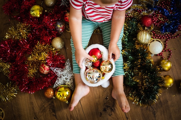 Kind sitzt auf dem Holzboden und hält die Weihnachtskugeln in der Tasche. Weihnachtsdekorationen.