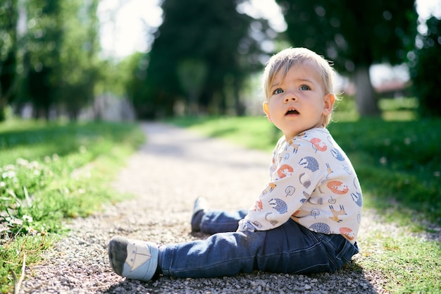 Kind sitzt auf dem Weg im Park und dreht den Kopf zur Seite