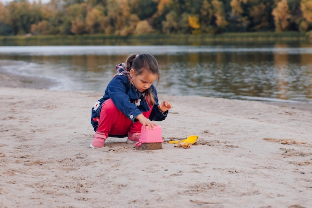 Kind spielt am Strand kleines Mädchen in blauer Jacke und rosa Hose macht Sandkuchen am Flussba...