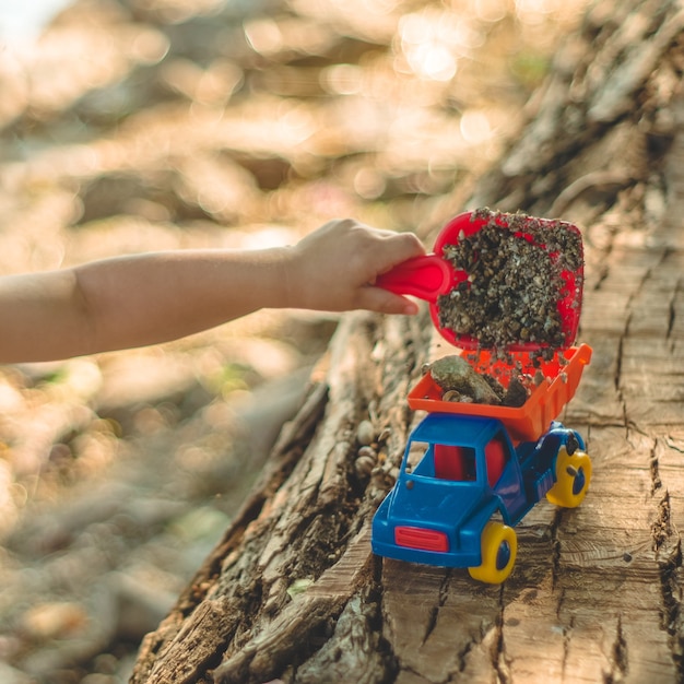 Kind spielt im Freien. Kind, wir gießen den Sand in den roten Lastwagen. Kinder Straßenspiele. Ein Junge spielt mit einer Maschine auf dem großen Baumstamm