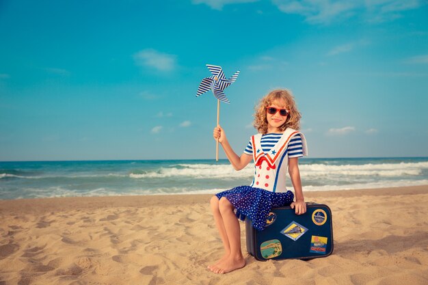 Kind spielt mit einem Windrad und sitzt auf einem Koffer am Strand gegen Meer- und Himmelshintergrund