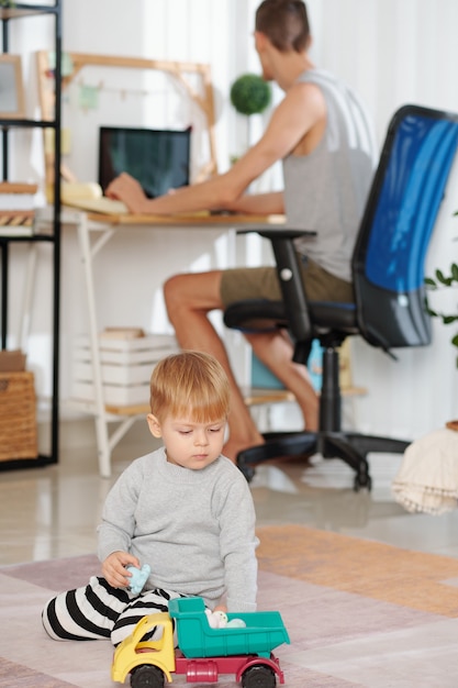 Kind spielt mit Spielzeug mit seinem Vater, der zu Hause am Computer im Hintergrund arbeitet