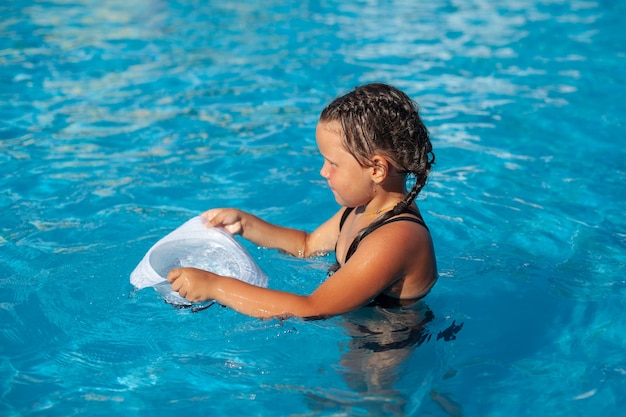 Kind steht im Pool kleines gebräuntes Mädchen mit geflochtenen Zöpfen auf dem Kopf und benetzt weiße Panama...