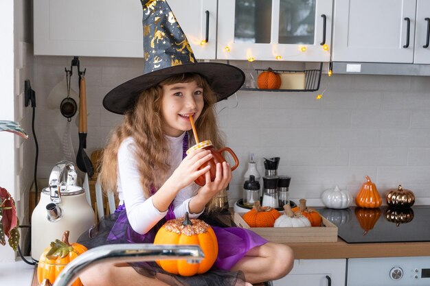 Kind trinkt Kürbissaft Gemüse-Smoothie oder Cocktail aus einem Becher mit Strohhalm Küche dekoriert für Halloween Mädchen in einem Hexenkostüm Herbst gemütlich zu Hause