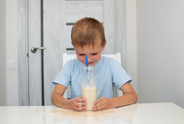 Kind trinkt zu Hause köstliche Schokoladenmilch. Niedlicher kleiner Junge trinkt ein Glas Milch und bläst Milchblasen, während er am Tisch sitzt