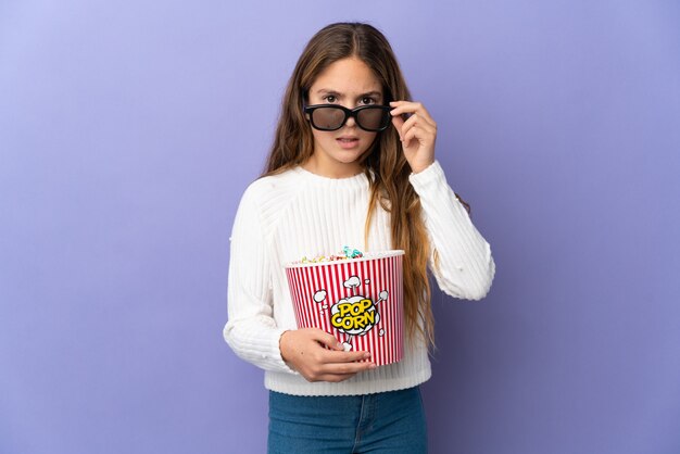 Kind über isolierte lila Wand überrascht mit 3D-Brille und hält einen großen Eimer Popcorn