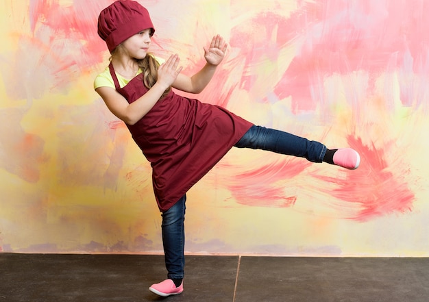 Foto kind und glückliche kindheit. kinderkoch in karate-pose an bunter wand. karriere, uniform, traum, beruf und berufskonzept. kochen, spielen, sport, fitness, energie. mädchen in kochmütze und schürze.