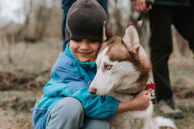 Kind und sein Freund Husky sibirischer Hund Porträt kleiner Junge umarmt süßes weißes braunes Säugetiertierhaustier von einem Jahr alt mit blauen Augen im Herbst rustikaler und ländlicher Naturwald