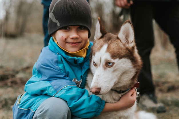 Kind und sein Freund Husky sibirischer Hund Porträt kleiner Junge umarmt süßes weißes braunes Säugetiertierhaustier von einem Jahr alt mit blauen Augen im Herbst rustikaler und ländlicher Naturwald