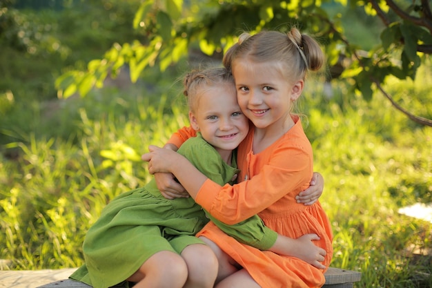 Kinder blonde Mädchen in einem gelben und grünen Kleid umarmen sich vor dem Hintergrund eines Freundesgartens