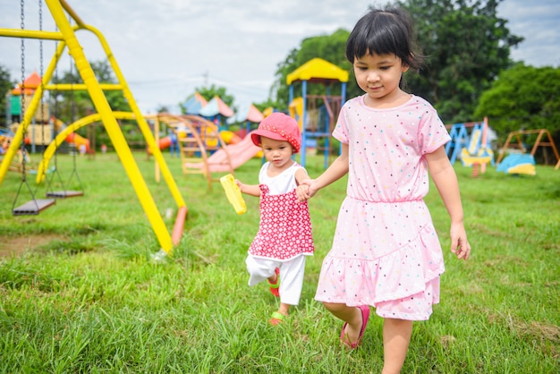 Kinder, die das kleine Mädchen des Spaßes hält Hand zusammen mit der Liebe spielt die äußeren asiatischen Kinder glücklich im Gartenpark mit Spielplatz haben