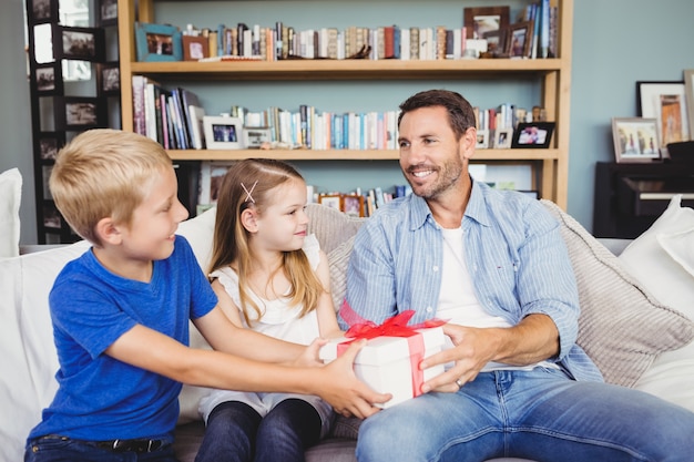 Kinder, die dem Vater Geschenkbox beim Sitzen auf Sofa geben