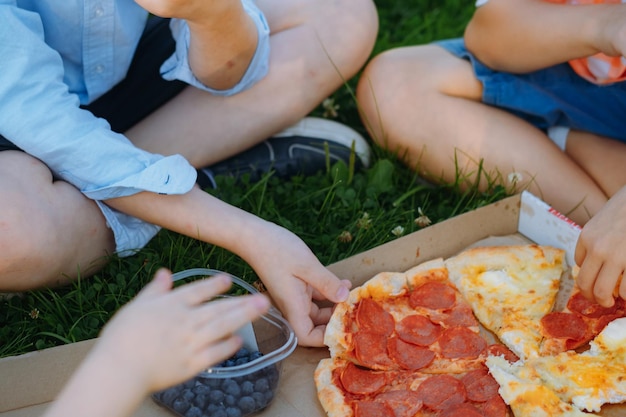 Kinder, die nach der Schule auf Gras zu Mittag essen. Kinderhände greifen nach Pizzastücken. Bild mit selektivem Fokus