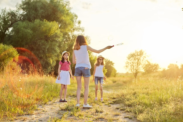 Kinder drei Mädchen spielen mit Seifenblasen, Teenager bläst jüngere Blasen fangen. Schöne Naturlandschaft, Sommerwiesenhintergrund, glückliche Kindheit