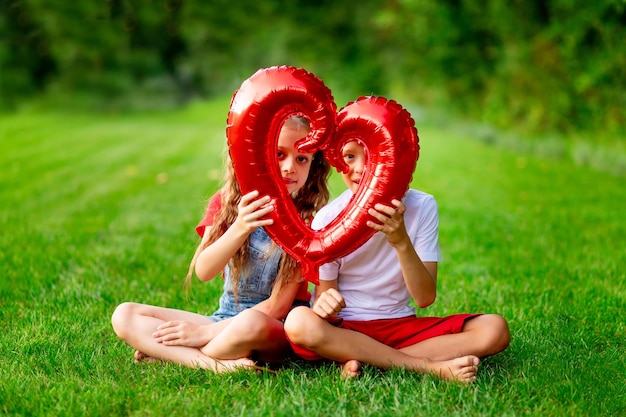 Kinder, ein Mädchen und ein Junge, blond mit einem großen roten Herzen im Sommer auf dem Rasen auf dem grünen Gras, haben Spaß und freuen sich über das Konzept des Valentinstags Platz für Text