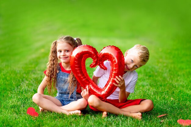 Kinder ein Mädchen und ein Junge blonde mit einem großen roten Herz im Sommer auf dem Rasen auf dem grünen Gras haben Spaß und freuen sich über das Konzept des Valentinstags Raum für Text