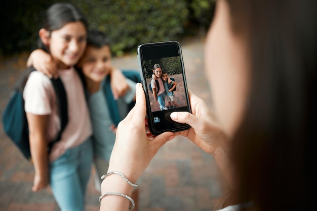 Kinder erster Schultag und Mutter fotografieren mit einem Telefon ihrer süßen Kinder Nahaufnahme des Bildschirmbildes von Bruder und Schwester, die sich umarmen, während sie morgens draußen für ihre Mutter posieren