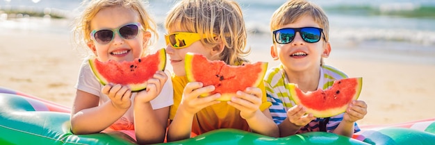 Kinder essen Wassermelone am Strand im langen Sonnenbrillen-Banner-Format